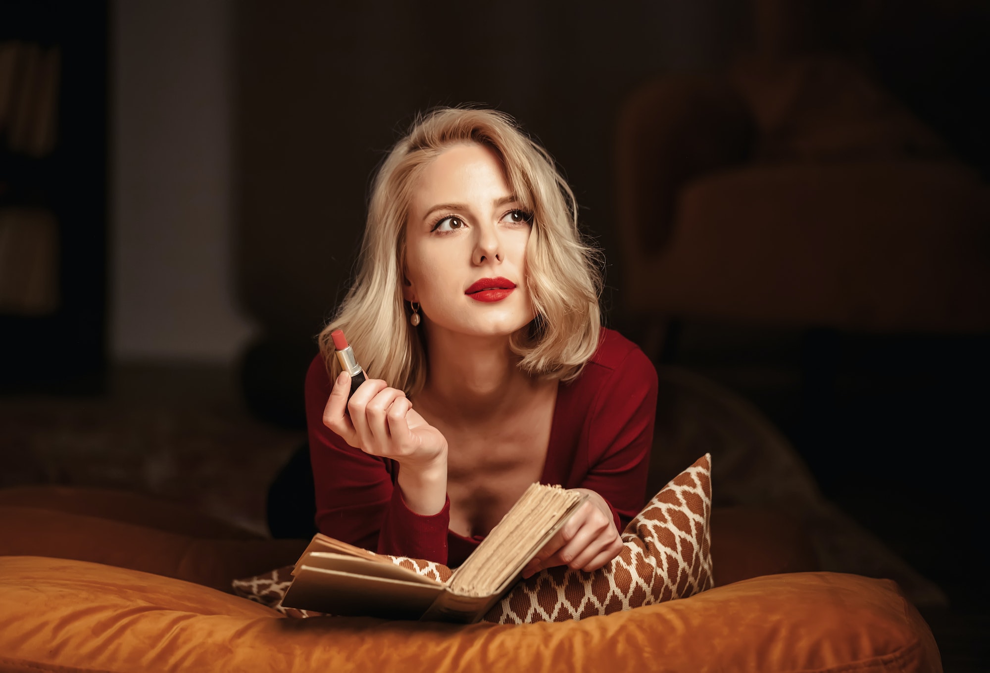 Beautiful blond girl with lipstick and book lies in pillows