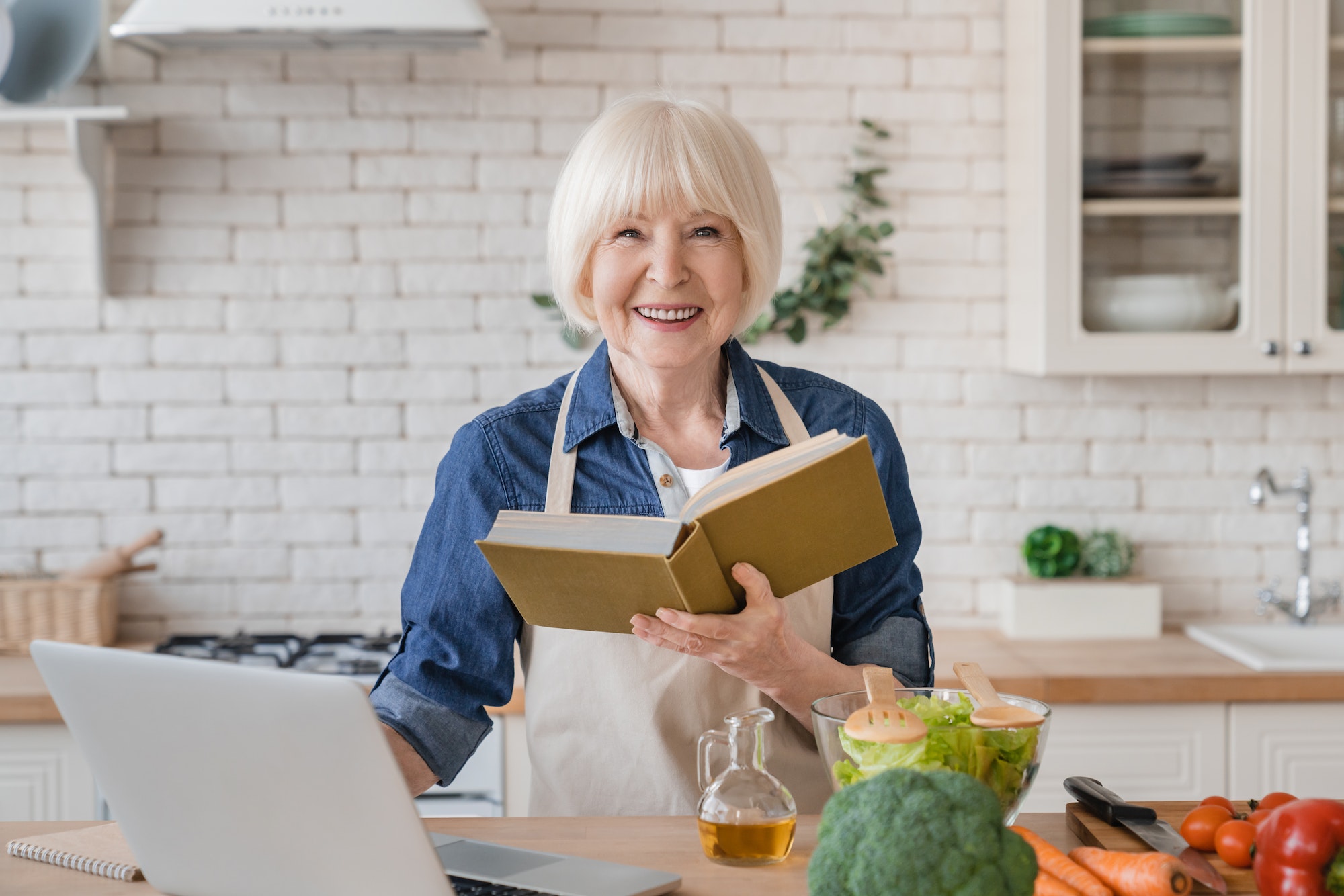 Caucasian aged old senior woman grandmother in apron reading a book of recipes, cooking meal